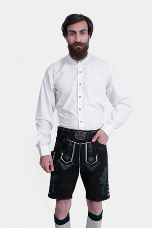 Full-body view of the same man wearing the white shirt and black lederhosen with green embroidery. He also has gray and green knee-high socks and black shoes, standing against a plain white background.