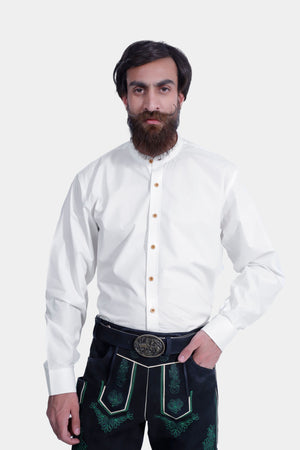 Close-up of the man wearing the white shirt and black lederhosen. The intricate green embroidery and decorative belt buckle are clearly visible. He stands against a plain white background with one hand in his pocket.