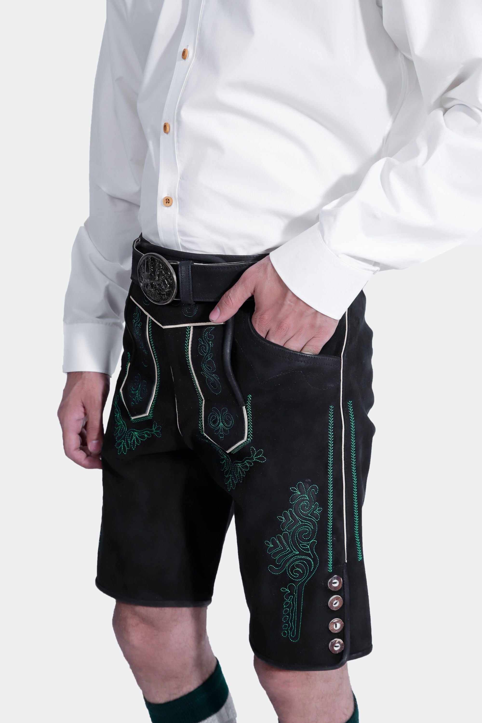 A side view close-up of the man's hand resting in the pocket of the black lederhosen, highlighting the green embroidery and side buttons. He is wearing a white button-up shirt, and the background is plain white.