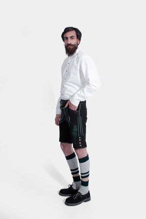 Side view of a man in a white shirt and black lederhosen with green embroidery, one hand in pocket, against a white background.