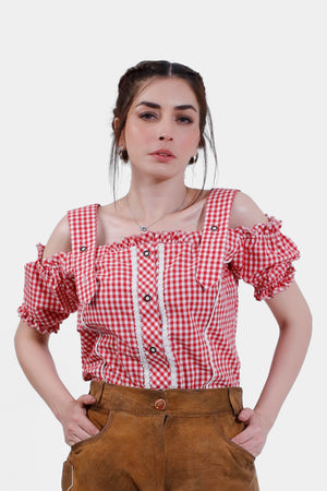 A woman wearing a red checkered off-shoulder traditional blouse with ruffled sleeves and decorative buttons. She pairs the blouse with tan leather lederhosen, standing confidently with her hands in her pockets. Her hair is styled in a braided updo, giving her a classic and elegant look.