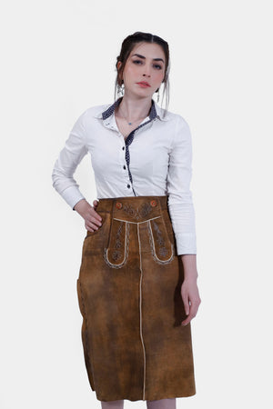 Model wearing a traditional brown leather Nena Lederhosen Skirt with white embroidery, buttons, and side pockets, paired with a white blouse with a polka-dotted inner collar
