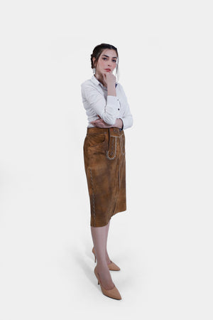 Full body side profile view women wearing a traditional brown leather Nena Lederhosen Skirt with white embroidery, buttons, and side pockets, paired with a white blouse with a polka-dotted inner collar