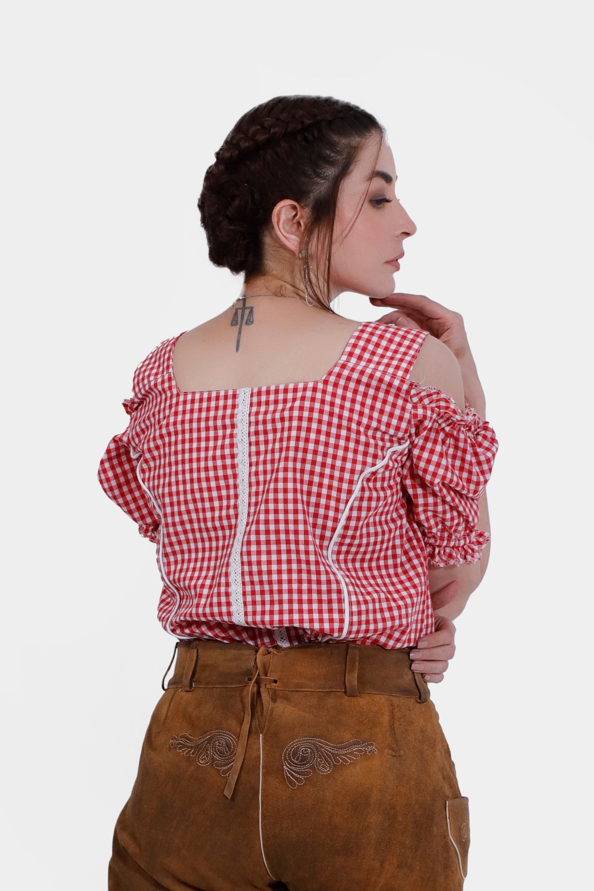 woman is shown from the back wearing a red checkered off-shoulder traditional blouse with ruffled sleeves and a lace trim down the back. She pairs the blouse with tan leather lederhosen, showcasing intricate embroidery on the back.