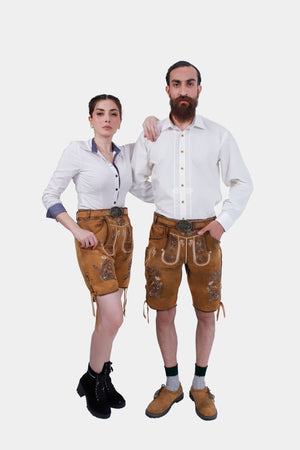A couple dressed in matching Neuschwanstein Couple Outfits. The woman is wearing a crisp white blouse paired with golden-brown leather shorts featuring intricate embroidery and black lace-up boots. The man is wearing a white button-down shirt with golden-brown leather shorts that have similar detailed embroidery, paired with brown shoes and green knee-high socks.