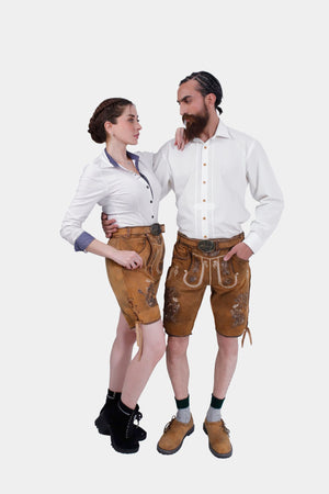 The couple posing in their Neuschwanstein Couple Outfits. The woman has her arm around the man's shoulder, showcasing her white blouse and golden-brown leather shorts with intricate embroidery. The man complements her look with a white button-down shirt and matching golden-brown leather shorts, finishing the outfit with brown shoes and green knee-high socks.