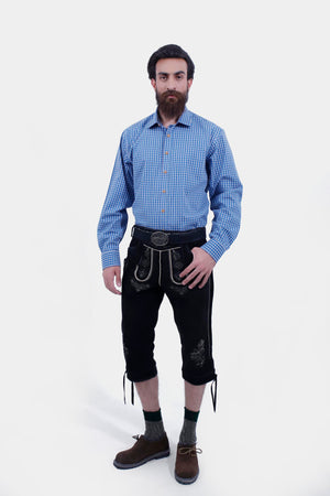 A man with a beard and mustache wearing a blue checkered shirt paired with black Bundhosen, standing with his hands in his pockets. The Bundhosen are decorated with intricate embroidery.