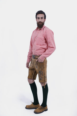 A man with a full beard wearing traditional Bavarian clothing, featuring a red and white checkered shirt and tan leather lederhosen with brown embroidery, paired with green knee-high socks and brown shoes, standing with one hand in his pocket.