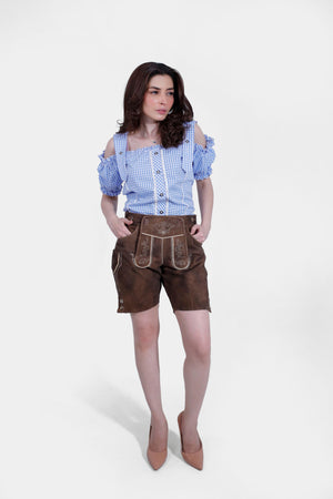 A woman wearing the Paula Beer Lederhosen Outfit, featuring high-quality leather shorts with intricate embroidery and a blue and white checkered off-shoulder Bavarian blouse. She stands confidently with her hands in the pockets of her lederhosen.
