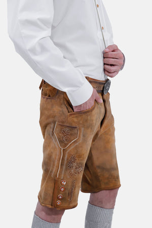 Side view of a man with a beard wearing a white dress shirt, with his hand in the pocket of his tan leather lederhosen shorts, highlighting the intricate embroidery and side button details, and grey knee-high socks.