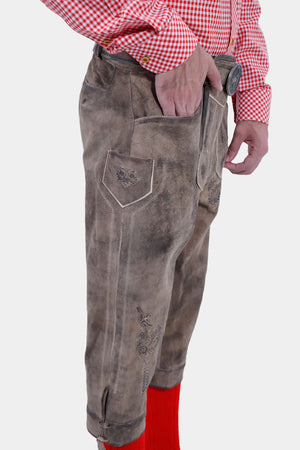 Side view of a man with a beard wearing a red and white checkered shirt, with his hand in the pocket of his light brown leather bundhosen, highlighting the intricate embroidery and pocket design, and bright red knee-high socks.