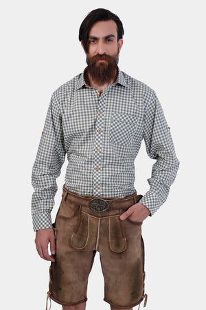 A man with a full beard wearing a gray and white checkered shirt and tan leather lederhosen with green embroidery, standing with one hand in his pocket, showcasing the detailed design of the lederhosen.