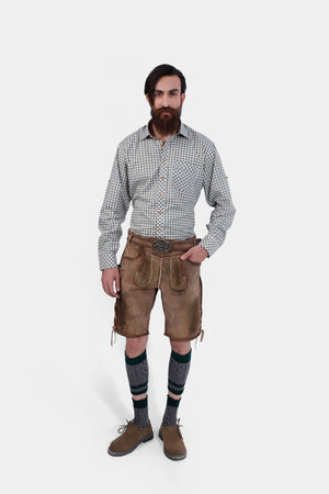 A front view of a man with a full beard wearing a gray and white checkered shirt and tan leather lederhosen with green embroidery, standing with both hands on his hips, showcasing the back design of the lederhosen.
