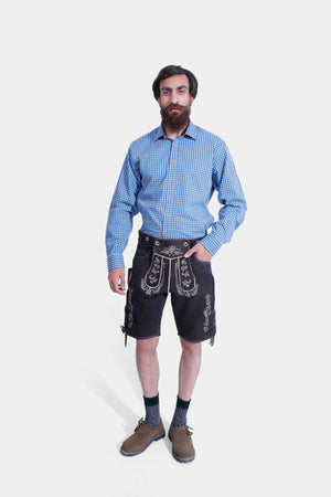 Man standing in a Gert Fröbe Lederhosen Outfit, including black leather shorts with white embroidery, a blue checkered shirt, and brown Bavarian shoes."