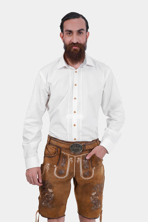 A man with a beard in a white dress shirt and brown lederhosen with detailed embroidery. He is standing facing the camera with a neutral expression, his left hand in his pocket.