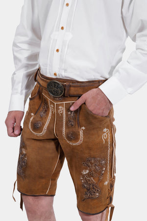 Close-up view of the brown lederhosen, showcasing the detailed embroidery on the front and sides. The man is wearing a white dress shirt and has his right hand in his pocket.