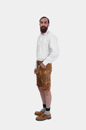 Side profile of a man wearing a white dress shirt and brown lederhosen with embroidery. He is standing with his left hand in his pocket and right hand on his waist. embroidery on the lederhosen. He has his left hand in his pocket and is looking slightly towards the camera.