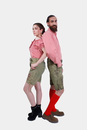 A couple standing back-to-back in their traditional Volksfest outfits. The woman wears a red checkered off-shoulder top and embroidered green leather shorts with black lace-up boots. The man is in a matching red checkered shirt and green leather shorts with detailed embroidery, paired with red knee-high socks and brown shoes.