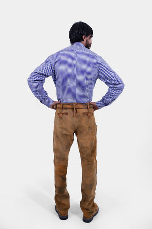 A full-length shot of a bearded man in a blue and white checkered shirt and brown leather long lederhosen, standing with his hands on his hips. The back view highlights the fit and details of the lederhosen.