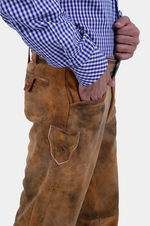 Close-up of a bearded man wearing a blue and white checkered shirt and brown leather long lederhosen. He has one hand in his pocket, highlighting the intricate details and texture of the lederhosen.