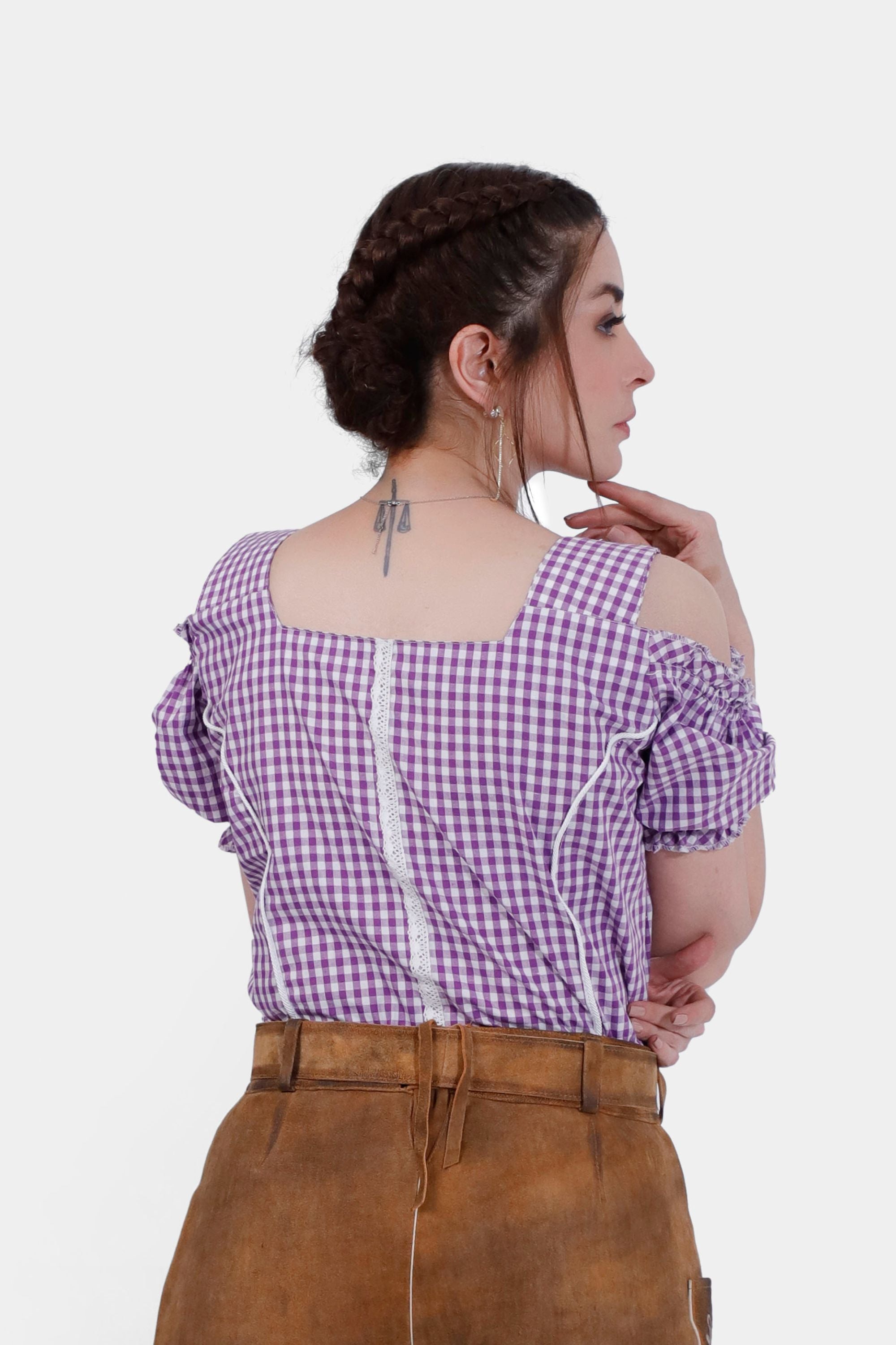 Woman wearing the Rihanna Traditional Blouse in purple checkered pattern, back view. The blouse features off-the-shoulder sleeves with ruffled edges, lace detailing along the back seam, and is paired with traditional leather trousers. The intricate design elements highlight the classic Trachten style.