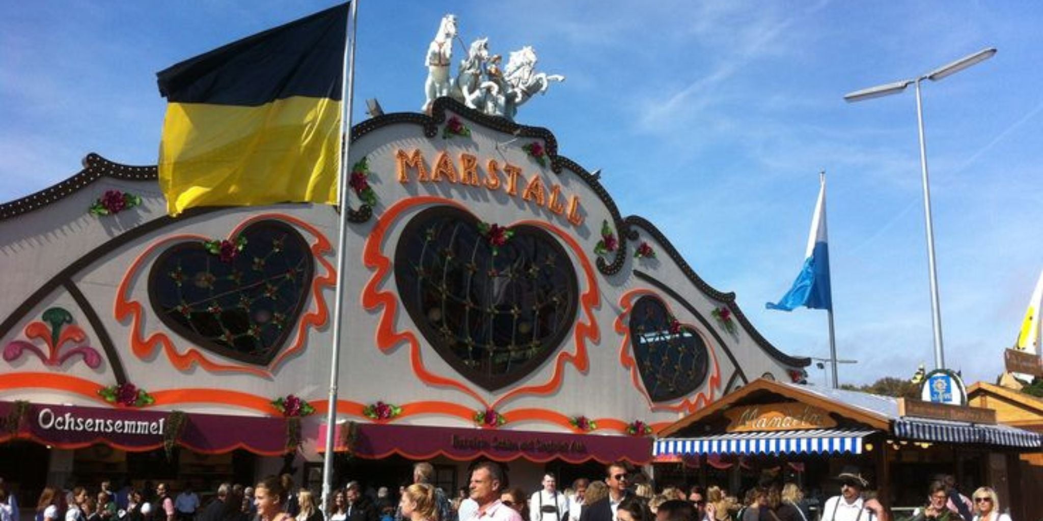Front view of the Marstall Festzelt tent at Oktoberfest Munich with heart-shaped windows, colorful decorations, and crowds of people in traditional Bavarian outfits