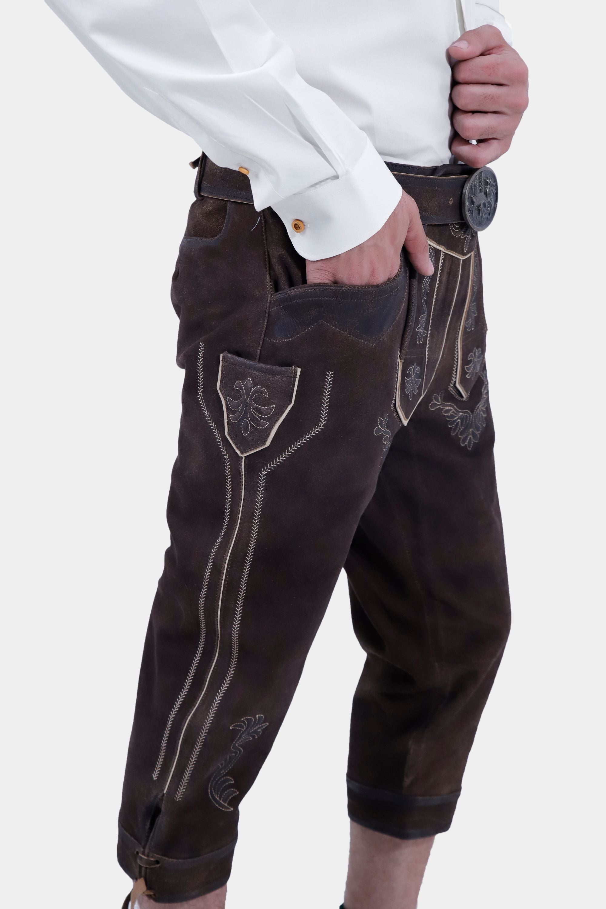 A man in a white shirt is posing in traditional dark brown leather Bundhosen. The outfit is accessorized with a belt and traditional Bavarian shoes and socks. The side view highlights the intricate embroidery on the Bundhosen.