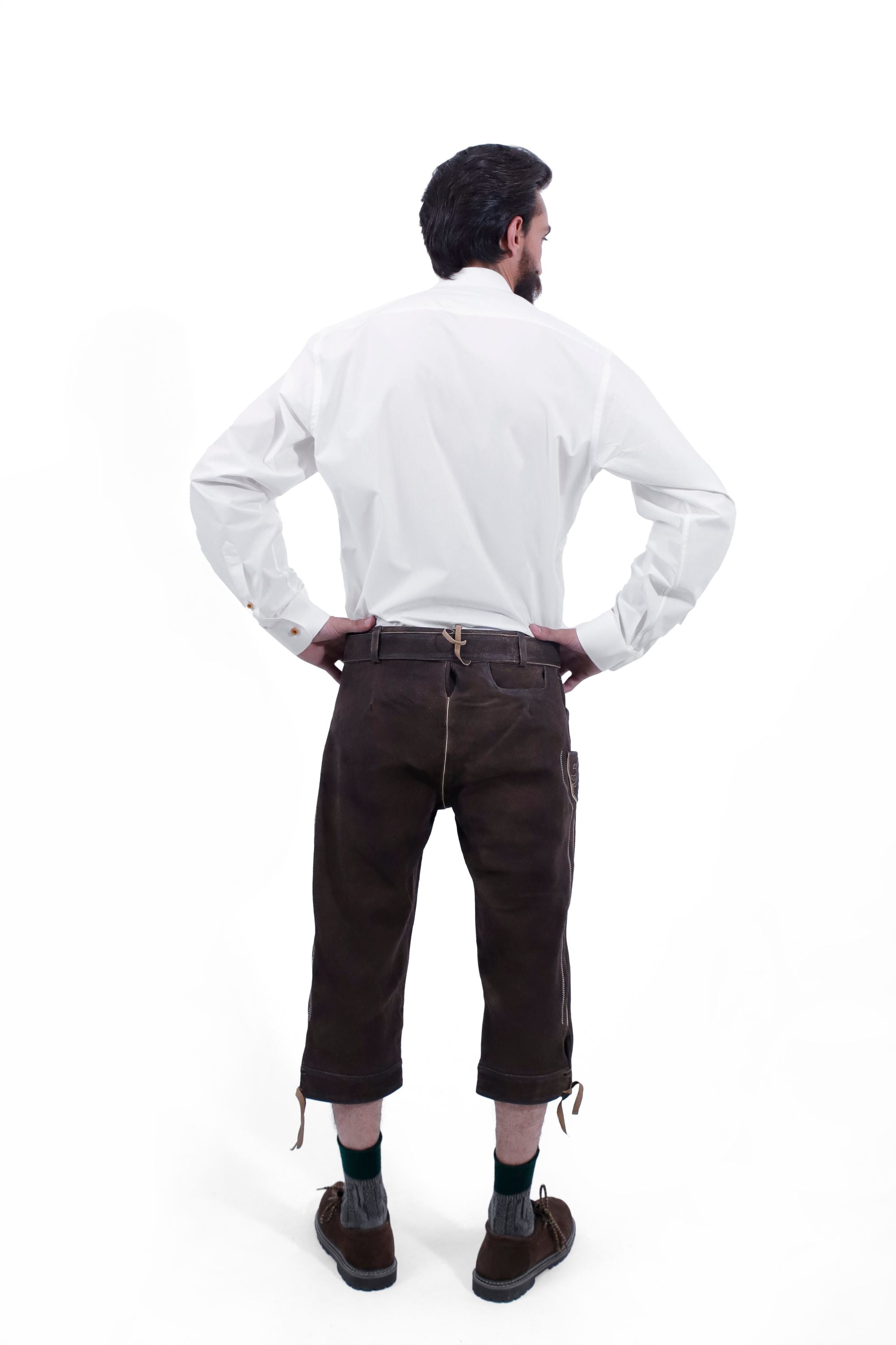Rear view of a man wearing traditional dark brown leather Bundhosen and a white shirt. The image showcases the back details of the Bundhosen, including the tie straps and embroidery on the back pocket. The man is also wearing traditional Bavarian socks and shoes.