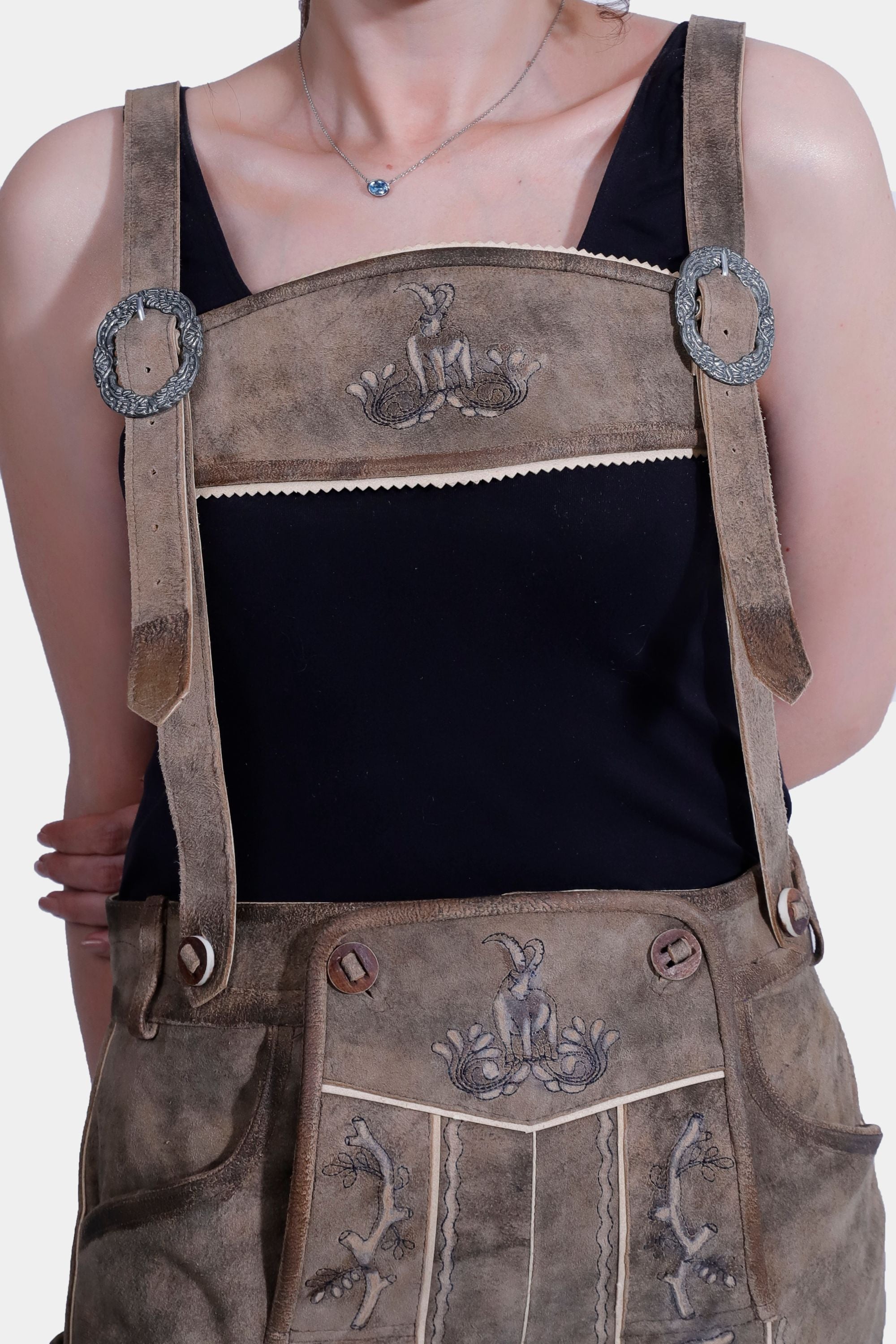 Close-up of a woman wearing traditional Bavarian Lederhosen with suspenders, featuring detailed alpine animal embroidery. The Lederhosen are light brown, and the background is plain white.