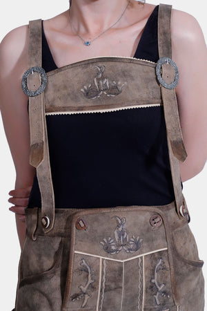 Close-up of a woman wearing traditional Bavarian Lederhosen with suspenders, featuring detailed alpine animal embroidery. The Lederhosen are light brown, and the background is plain white.