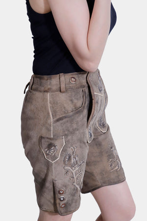 Side profile of a woman posing with one hand on her shoulder, wearing light brown Lederhosen with intricate embroidery. She is dressed in a black tank top, with a plain white background.