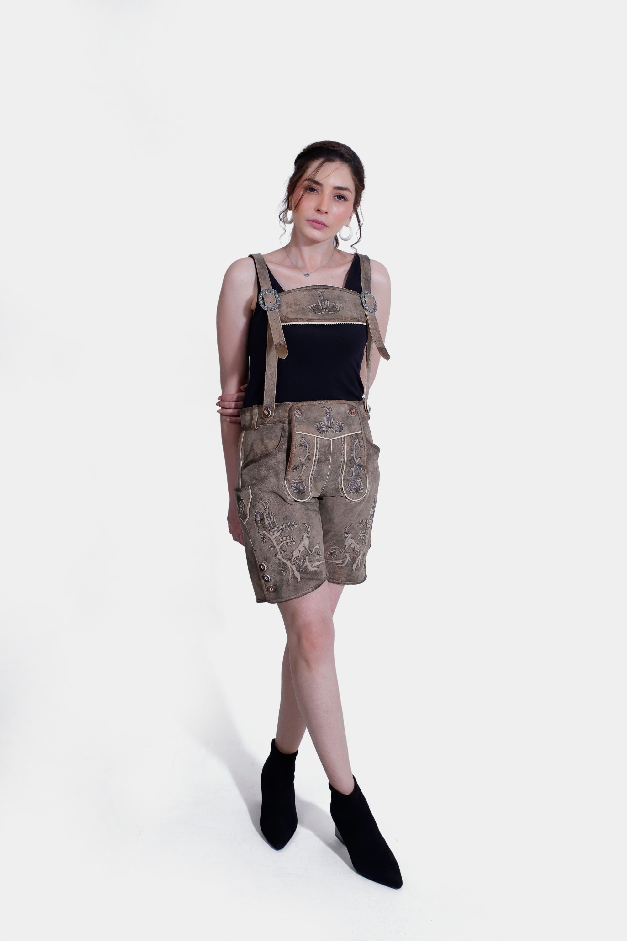 A full-body image of a woman standing in light brown Lederhosen with detailed embroidery, hands on her hips. She wears a black tank top, with a plain white background.