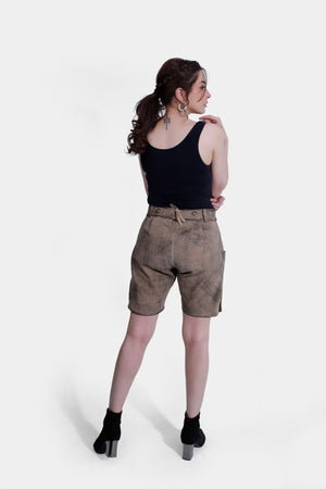 A full-back body image of a woman standing in Lederhosen with detailed embroidery, hands on her hips. She wears a black tank top, with a plain white background.