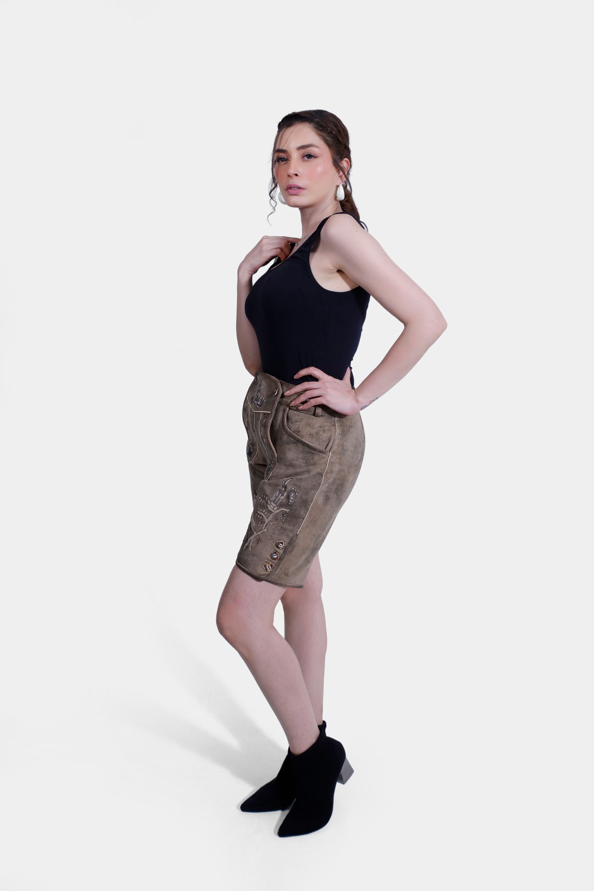 A full-body side profile of a woman in light brown Lederhosen with detailed embroidery, wearing a black tank top and black ankle boots. The background is plain white.