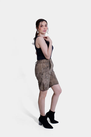 Side profile of a woman in light brown Lederhosen with intricate embroidery, paired with a black tank top and black ankle boots. The background is plain white.