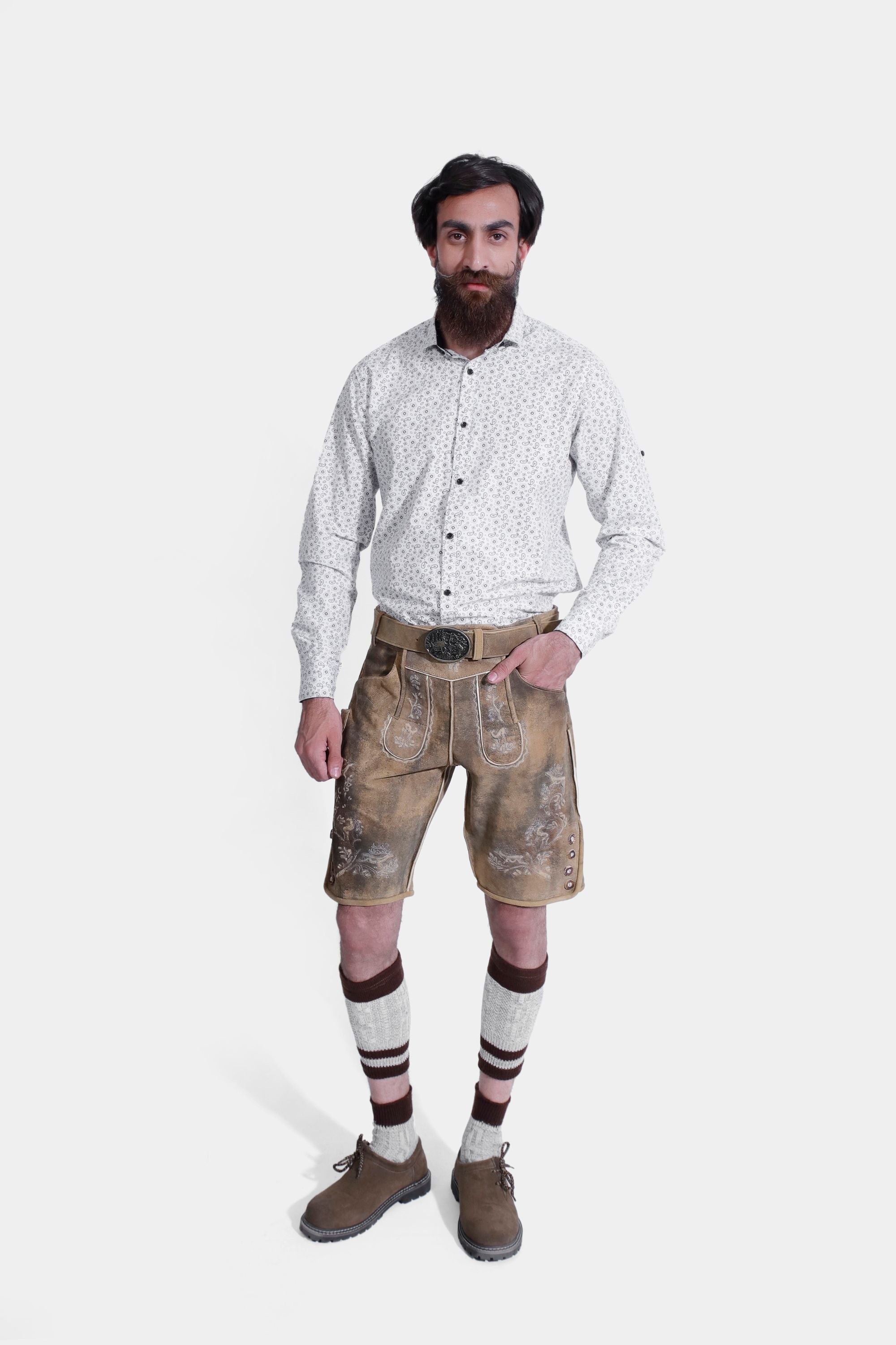 The man stands confidently with one hand in his pocket, wearing the lederhosen and the patterned white shirt. The image provides a clear view of the embroidery on the lederhosen and the ornate belt buckle.