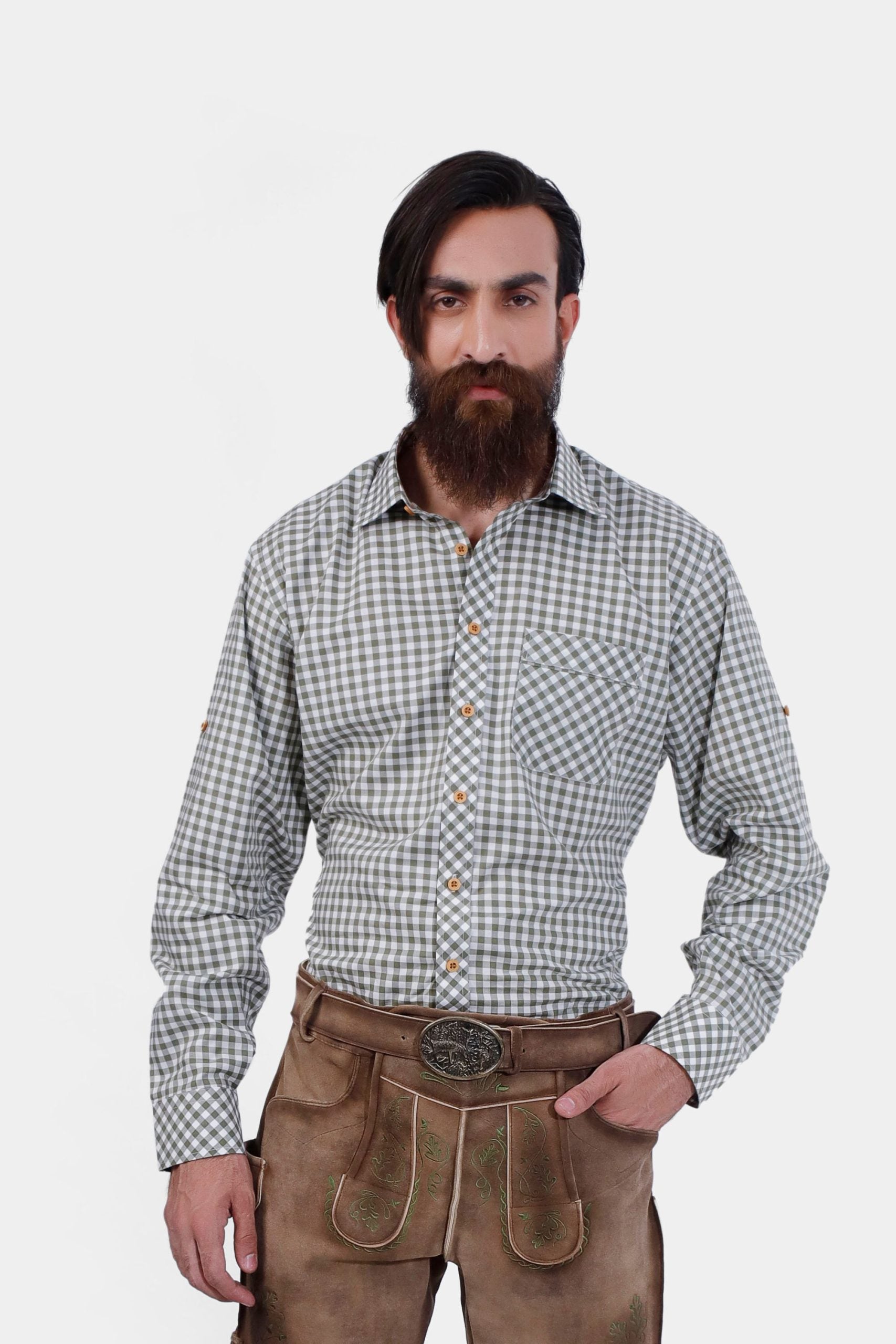 A bearded man wearing the André Lederhosen Shirt, featuring a green and white checkered pattern. He is paired with traditional beige lederhosen embroidered with green detailing. The man stands confidently with one hand in his pocket against a plain white background, showcasing the classic Bavarian attire.