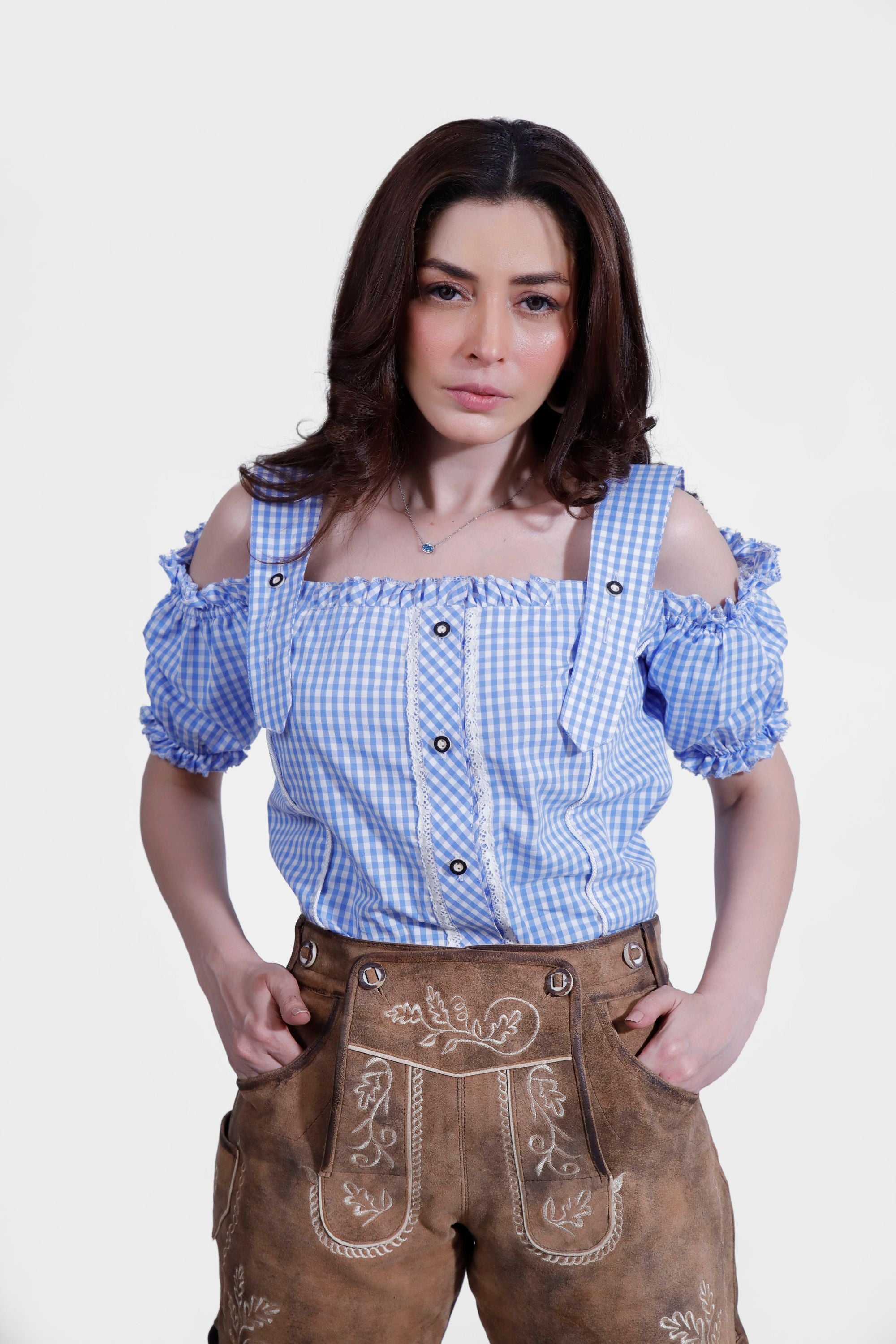 Close-up of a woman in a blue and white checkered off-shoulder blouse and brown short lederhosen. She has her hands in her pockets, highlighting the intricate embroidery and design of the lederhosen.