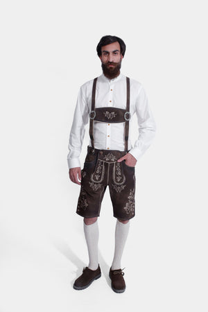 A man with a beard stands against a white background, wearing the "Angry Stag Eibsee Lederhosen." These traditional dark brown Lederhosen are knee-length and feature intricate light-colored embroidery on the bib, side panels, and around the leg openings. Complemented by a crisp white shirt, light grey knee-high socks, and dark brown shoes, his attire is a classic example of authentic Alpine fashion.