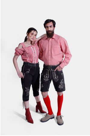 A couple dressed in matching Bamberger Couple Outfits. The woman is wearing a red checkered off-shoulder blouse paired with elegant black knee-length lederhosen adorned with white embroidery and stylish red ankle boots. The man is wearing a red checkered shirt with black lederhosen shorts, also decorated with white embroidery, beige shoes, and bright red knee-high socks.