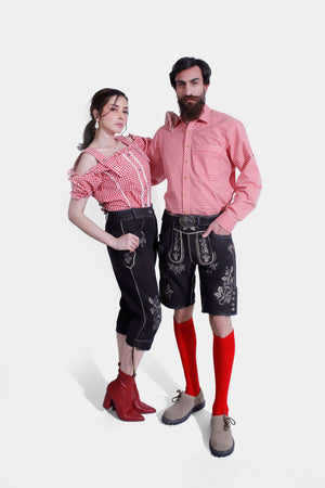 The couple posing in their Bamberger Couple Outfits. The woman has her arm around the man's shoulder, showcasing her red checkered off-shoulder blouse and embroidered black knee-length lederhosen. The man complements her look with a matching red checkered shirt and black lederhosen shorts with detailed white embroidery.