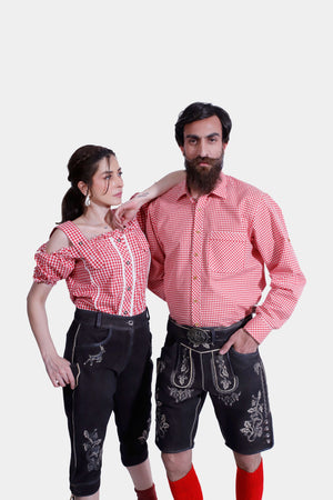 A couple dressed in matching Bamberger Couple Outfits. The woman is wearing a red checkered off-shoulder blouse paired with elegant black knee-length lederhosen adorned with white embroidery and stylish red ankle boots. The man is wearing a red checkered shirt with black lederhosen shorts, also decorated with white embroidery, beige shoes, and bright red knee-high socks.