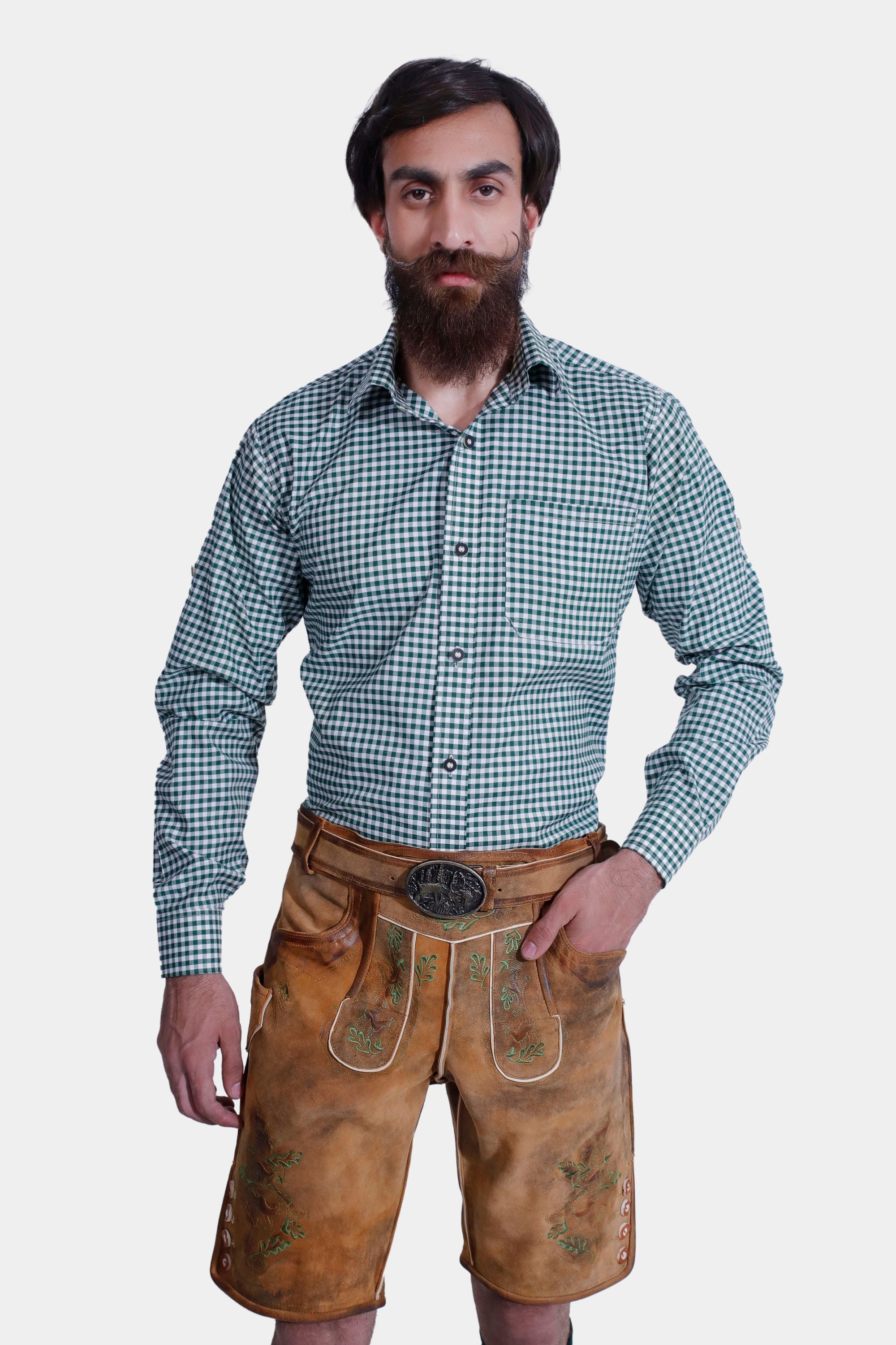 A man with a full beard and mustache, dressed in a green checkered shirt paired with tan leather lederhosen. The lederhosen feature intricate green embroidery and a matching belt with an ornate buckle. He stands with one hand in his pocket.