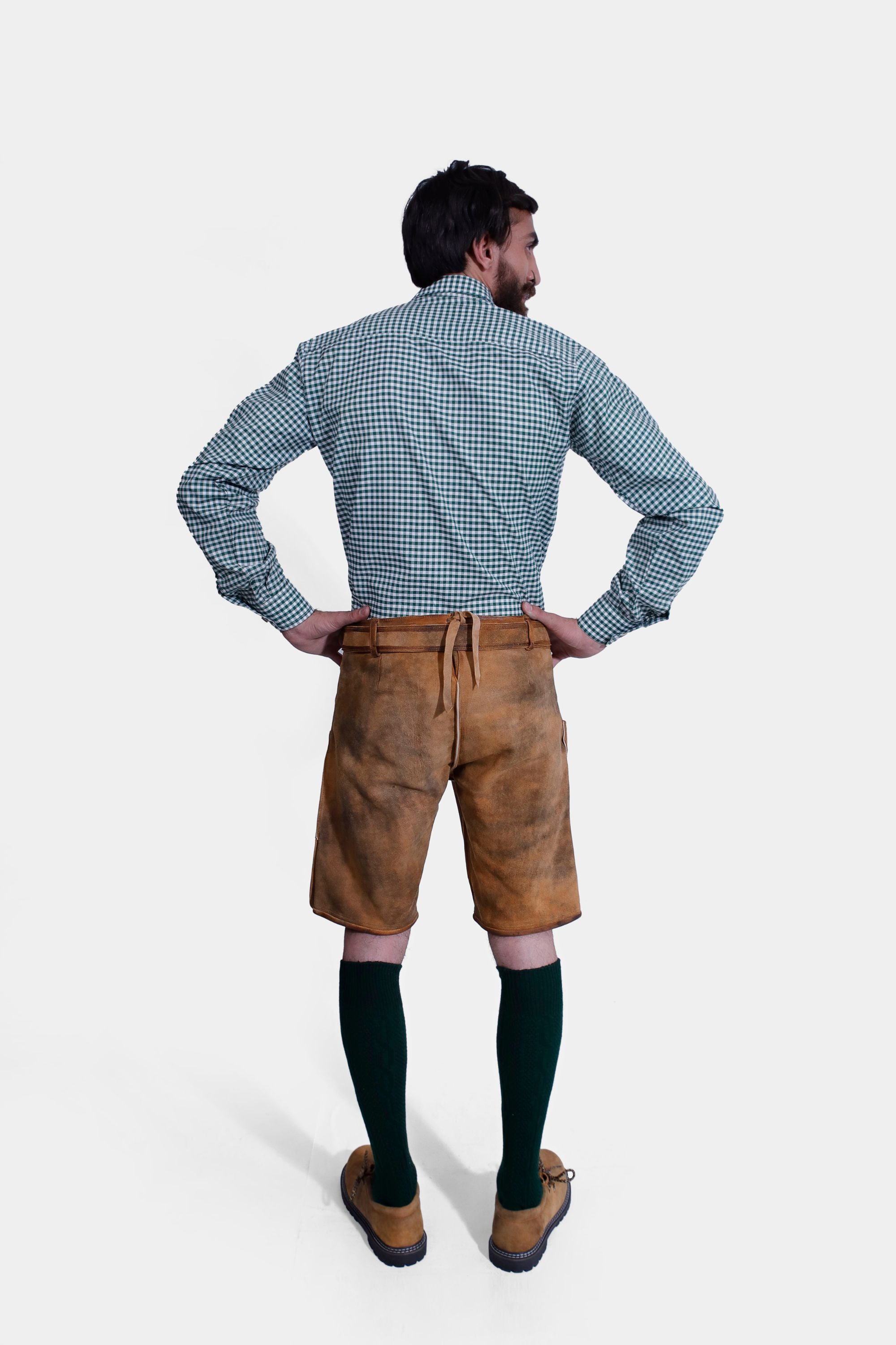 A rear view of the man, showcasing the back of the lederhosen and the green checkered shirt. The lederhosen have a small pocket and an adjustable strap at the back for a perfect fit.