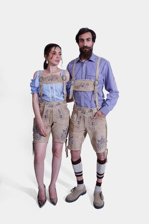 A couple dressed in matching Berliner Couple Outfits. The woman is wearing a blue checkered off-shoulder blouse paired with light tan leather shorts featuring intricate blue embroidery. The man is wearing a blue checkered shirt with light tan leather shorts, also adorned with blue embroidery, and beige shoes with traditional Bavarian socks.