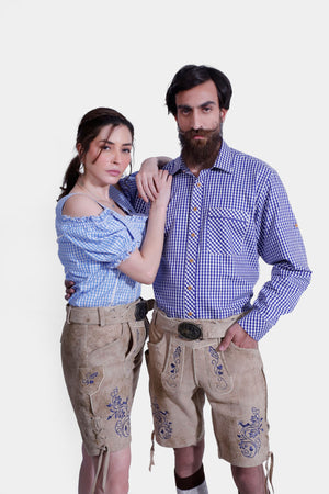 The couple posing in their Berliner Couple Outfits. The woman has her arm around the man's shoulder, showcasing her blue checkered off-shoulder blouse and embroidered light tan leather shorts. The man complements her look with a matching blue checkered shirt and embroidered light tan leather shorts.