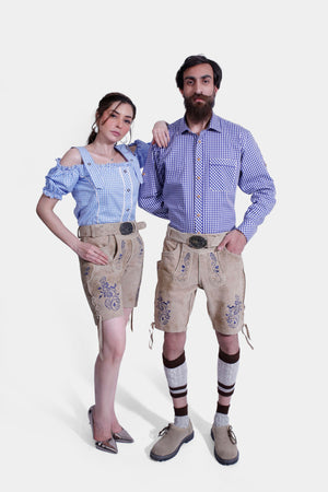 The couple standing back-to-back in their Berliner Couple Outfits. The woman wears a blue checkered off-shoulder blouse and light tan leather shorts with blue embroidery. The man is in a matching blue checkered shirt and light tan leather shorts with detailed blue embroidery, paired with beige shoes and traditional Bavarian socks.