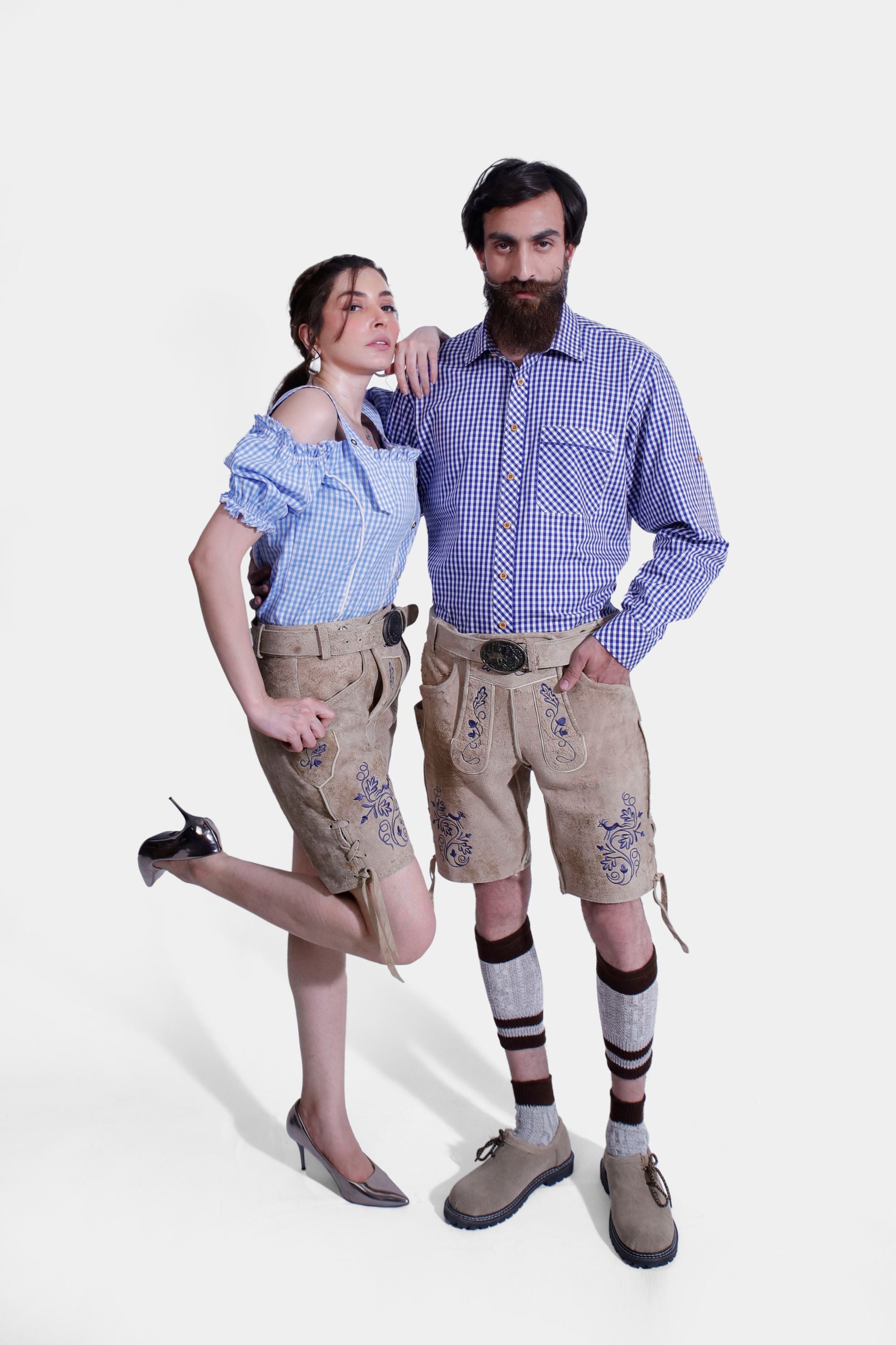 The couple in their Berliner Couple Outfits, standing side by side. The woman is wearing a blue checkered off-shoulder blouse with light tan leather shorts featuring blue embroidery and sleek, pointed high heels. The man is dressed in a matching blue checkered shirt and light tan leather shorts with intricate blue embroidery, beige shoes, and traditional Bavarian socks.