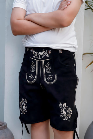 Close-up of a young boy in a white t-shirt wearing 'Bernd Leno Lederhosen' in black, showcasing detailed white embroidery on the bib and pockets, standing with arms crossed.