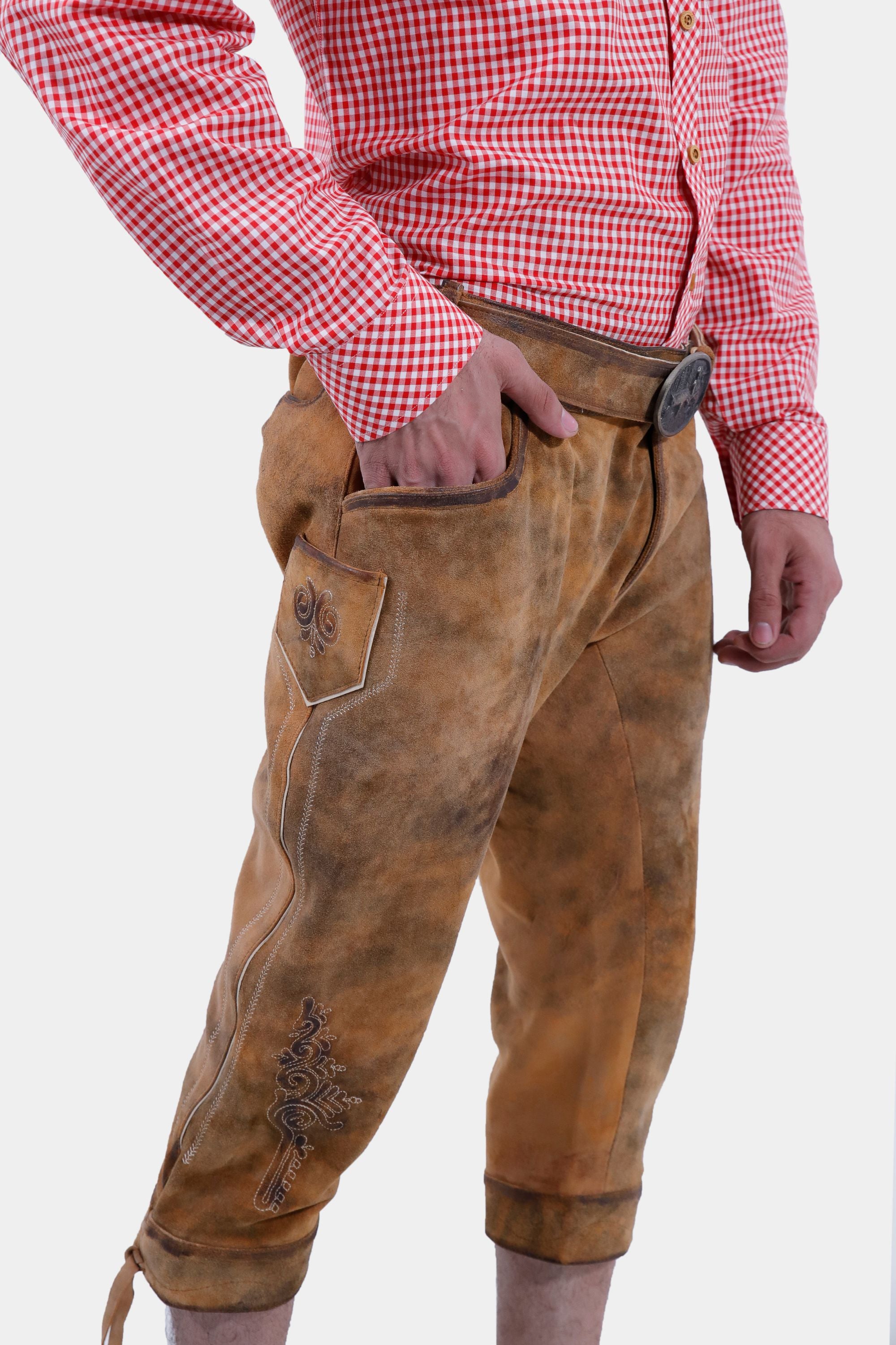 Close-up side view of a man wearing a red and white checkered shirt paired with brown leather Bundhosen. The image focuses on the detailed embroidery on the side of the pants and the rugged texture of the leather. His hand rests in the pocket, showcasing the functional design.